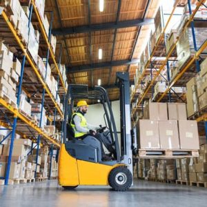 A man on a fork lift truck moving pallets in a warehouse surrounded by racking