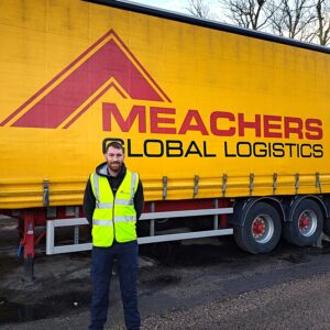 A man in high vis posing in front of a yellow and red Meachers truck celebrating his CPC pass